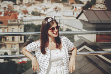 Fototapeta premium Beautiful stylish young woman in sunglasses, retro dress and headband relaxing on roof in european city street. Happy hipster girl smiling and enjoying summer time in Lviv. Copy space