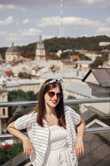 Beautiful stylish young woman in sunglasses, retro dress and headband relaxing on roof in european city street. Happy hipster girl smiling and enjoying summer time in Lviv. Copy space