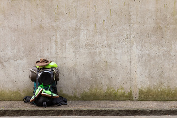 Hiking backpack with green towel, frying pan, sleeping bag, leather hat and green running shoes standing on a roadside in front of a gray wall