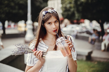 Beautiful stylish young woman holding drink and relaxing in city street. Happy hipster girl in retro dress and headband, holding plastic cup and straw, drinking lemonade.