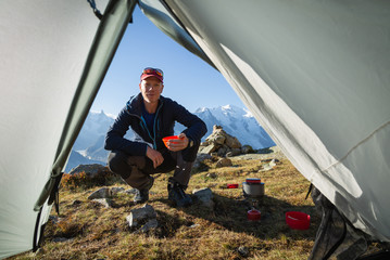 Hiker looking inside his tent and enjoying a cup of coffe at his mountain campsite.