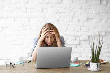 Frustrated young woman looking at laptop with stressed facial expression, keeping hands on head,...