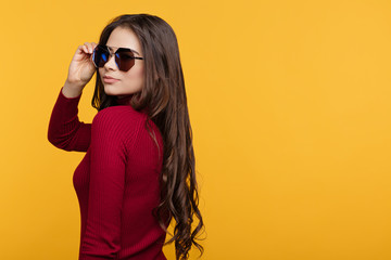 Pretty young lady in red dress and sun glasees is posing with hand on face on orange background.