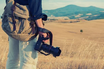 Tuinposter Man professional photographer with digital camera walking in tuscany fields. Photo Camera in male hands over summer landscape in Tuscany with fields, blue sky and moutains, Italy, Europe. © Khorzhevska