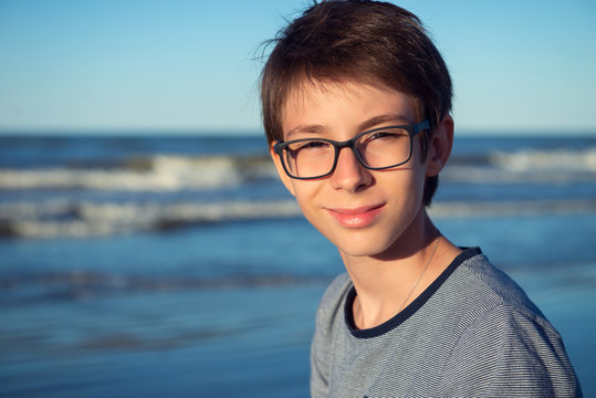 Young boy posing at the summer beach. Cute spectacled smiling happy 12 years old boy at seaside, looking at camera. Kid's outdoor portrait over seaside.