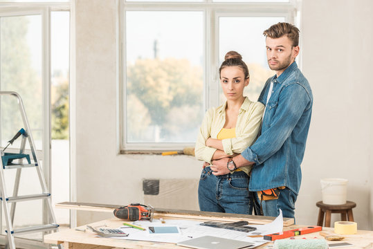 Serious Young Couple Embracing And Looking At Camera During House Repairment