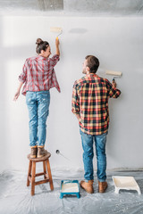 back view of happy young couple painting wall with paint rollers
