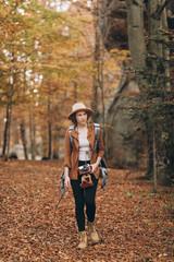Naklejka premium Attractive young woman in a stylish hat and travel bag on her shoulders, looking around at the charming autumn forest
