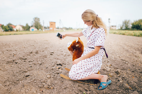 Nice looking beautiful blonde girl making selfie photo on smartphone. Young happy pretty female in stylish retro dress riding toy horse outdoor. Strange bizarre fashionable woman portrait.  Childhood.