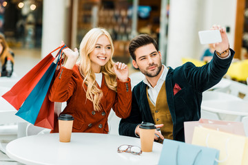 fashionable couple with shopping bags and coffee to go taking selfie on smartphone in shopping mall