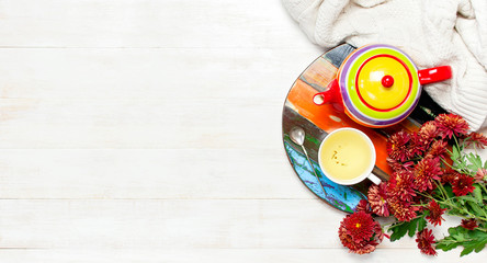Cup of hot herbal tea, bright colored teapot on wooden tray blackboard, knitted plaid or sweater red chrysanthemum flowers on light wooden background top view Copy Space Cozy morning breakfast at home