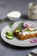Herring, red onions, cucumbers  and a slice of rye bread on a light grey plate.