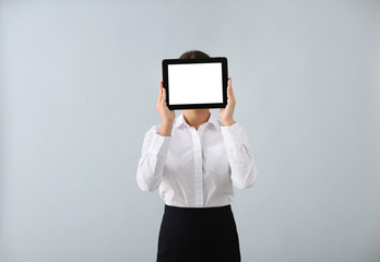 Beautiful young woman hiding face behind tablet computer on light background