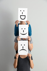 Young women hiding faces behind sheets of paper with drawn emoticons on light background