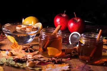 Dark wooden table with 2 glasses of tea with fall, autumn decoration and dark, black background. Colorful leaves, lemon, cinnamon, walnuts, apples, tea, walnut shells, hazelnuts, tea strainer, acorns.
