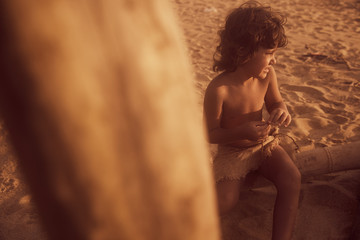 Mowgli boy with curly hair on the beach, sitting on big bamboo stick and playing with rope.