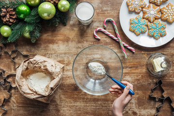 cropped shot of woman pouring flour into bowl while making dough for christmas cookies on wooden tabletop with decorative wreath