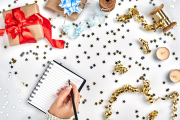 Festive decorations, delicate wavy ribbons, metallic star shaped confetti and notebook with wish list on white table, flat lay style. Christmas holidays decoration concept.
