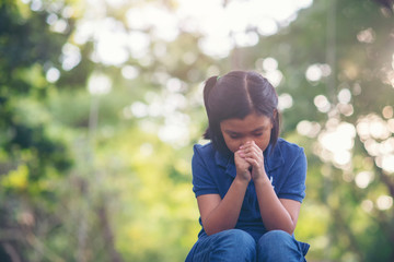 Asian child praying and hopeful for peace the world.Little kid sitting and hands in hands together in park,believes and faith in christian religion.Prayer and hopeful concept.