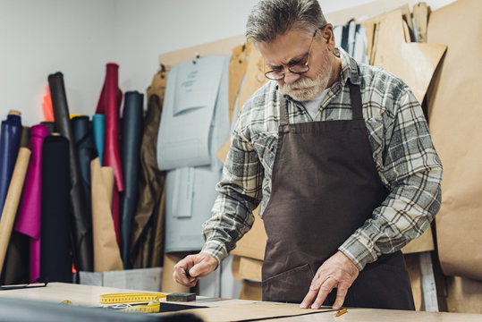 selective focus of mature leather handbag craftsman in apron and eyeglasses working at studio