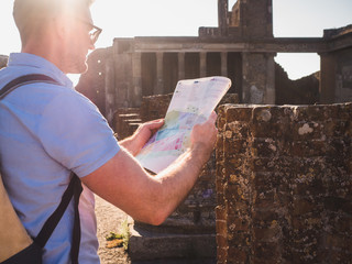 Handsome man in sunglasses looking at a map with landmarks against the backdrop of historic ruins, blue sky and the rays of the setting sun. Italy, Pompeii. Travel and vacation concept