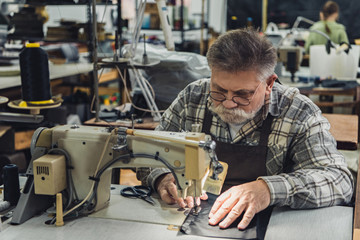 serious mature male tailor in apron and eyeglasses working on sewing machine at studio