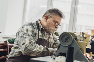 focused mature male tailor in apron and eyeglasses working on sewing machine at studio