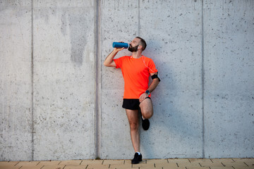 Fototapeta na wymiar Man resting from running and drinking water while standing against the wall. Healthy lifestyle concept.