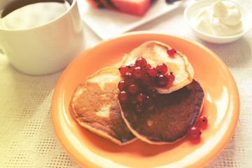 pancakes in orange plate and glass of sourcream with red berries, slice of watermelon, wooden table and white cloth.