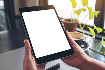 Mockup image of hands holding and using black tablet pc with blank white desktop screen with notebook, green leaves and coffee cup on wooden table