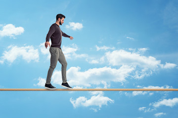 A casually dressed man with a beard walks carefully on a tight rope on a sky background. - obrazy, fototapety, plakaty