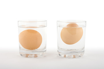 Two glasses of water with a fresh egg on the left and a rotten egg on the right side on white...