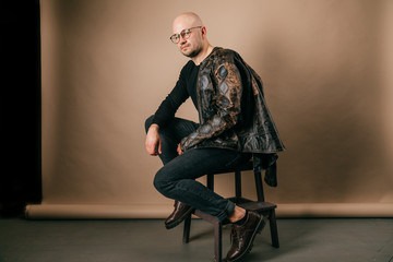 Lifestyle portrait of fashionable stylish bald man in glasses. Awesome man wearing biker leather jacket, black jeans, oxford shoes. Cool handsome well dressed boy on coffee paper studio background.