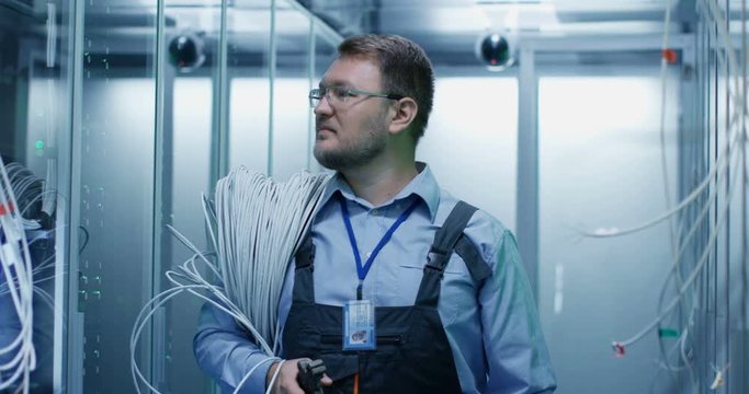 Portrait of adult bearded man technician standing in corridor with cable on his shoulders of server room in data center smiling at camera