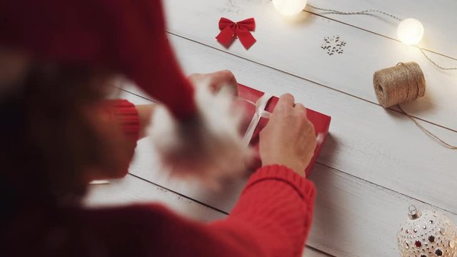 A woman in a red Santa Claus hat is tying a white ribbon on a red box with a gift against the background of white wooden boards, Christmas lights, Christmas toys, a skein. Play Christmas tunes. Bokeh