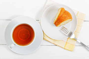 Saucer with a piece of homemade lemon cake and cup of tea on white wooden table. Top view.