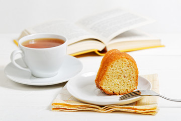 Piece of homemade lemon cake and cup of tea on white wooden table. Open book on blurred background. Shallow focus.