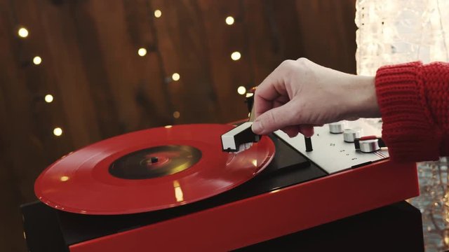 A young woman in a red knitted winter pullover puts a red vinyl record on a turntable, a stylus a needle and listens to music against the background of a glowing Christmas tree and a cozy New Year's