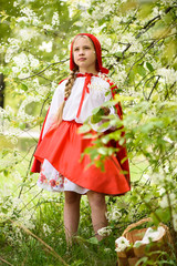 Smiling blond girl posing in a dress of little red riding hood near green trees