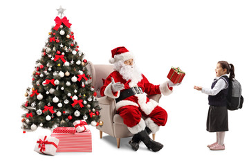 Santa Claus sitting in an armchair next to a Christmas tree and giving a present to a surprised little schoolgirl