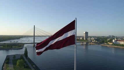 Aerial drone close-up shot rotating around a large flag of Latvia with skyline of Riga in background