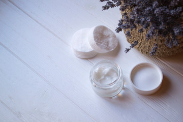 A jar with skin cream and lavender flowers on a white wooden background.
