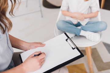 cropped shot of psychologist with clipboard sitting in front of little child in office