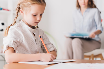 Naklejka na ściany i meble concentrated little child drawing while psychologist with clipboard sitting blurred on background