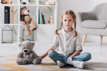 lonely little child sitting on floor with teddy bear and looking at camera