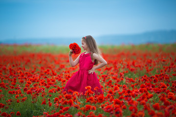Adorable blond girl dressed in vamp dark red stylish attractive dress posing on meadow of poppies field with mountains on background. Scene of cute teen holding bouquet of poppie in hands