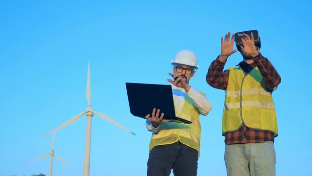 Energetics engineer in virtual reality glasses is being navigated by another specialist near windmill.
