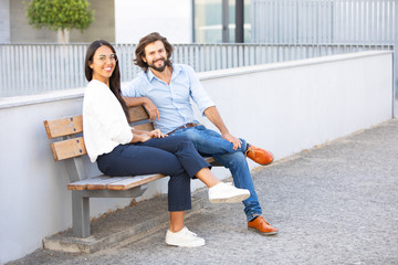 Smiling multiethnic couple sitting on bench near office building. Cheerful multicultural friends...