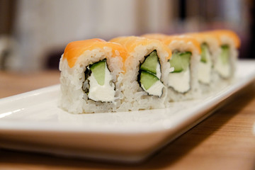 Horizontal shot of a close-up of rice rolls with salmon, cucumber and cream cheese on a white porcelain stretched rectangular dish on a wooden table in natural light colors.