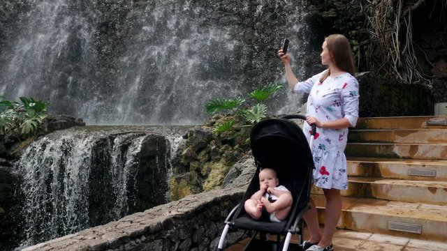 A young mother walking with her baby near the waterfall takes pictures on her smartphone for social networking and posting online while traveling with her family.